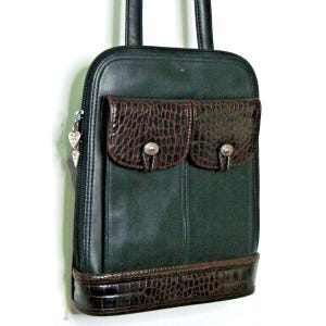 Puede incluir: Un bolso de mano de cuero verde oscuro con dos bolsillos marrones con textura de cocodrilo y una base a juego. El bolso tiene una cremallera y asas negras, y un dije plateado en forma de coraz&oacute;n. El bolso mide aproximadamente 30 cm de alto.