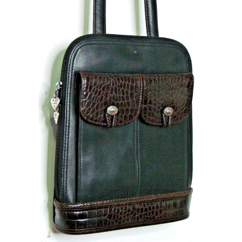 Puede incluir: Un bolso de mano de cuero verde oscuro con dos bolsillos marrones con textura de cocodrilo y una base a juego. El bolso tiene una cremallera y asas negras, y un dije plateado en forma de coraz&oacute;n. El bolso mide aproximadamente 30 cm de alto.