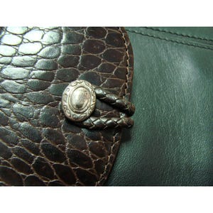 Puede incluir: Primer plano de una cartera de cuero marr&oacute;n con un patr&oacute;n texturizado similar a la piel de cocodrilo. La cartera presenta un bot&oacute;n ovalado plateado con un dise&ntilde;o intrincado y una correa de cuero trenzado.