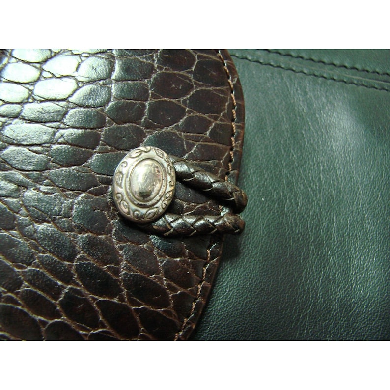 Puede incluir: Primer plano de una cartera de cuero marr&oacute;n con un patr&oacute;n texturizado similar a la piel de cocodrilo. La cartera presenta un bot&oacute;n ovalado plateado con un dise&ntilde;o intrincado y una correa de cuero trenzado.