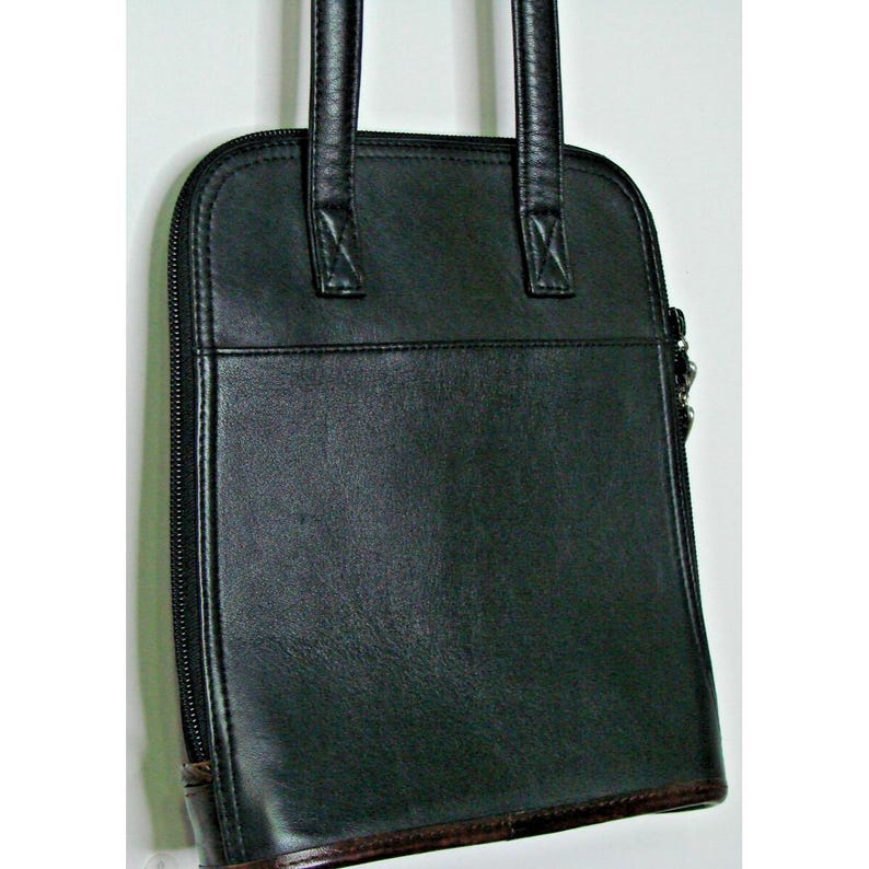 Puede incluir: Bolso tote de cuero negro con forma rectangular y cierre de cremallera. El bolso tiene dos asas y un ribete marr&oacute;n en la parte inferior. El bolso est&aacute; hecho de un material liso y brillante.