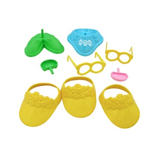 May include: A collection of colorful plastic toy accessories, including yellow visors with floral trim, yellow eyeglasses, a blue bib with a skull and crossbones design, and green and pink pieces. The items are arranged on a white background.