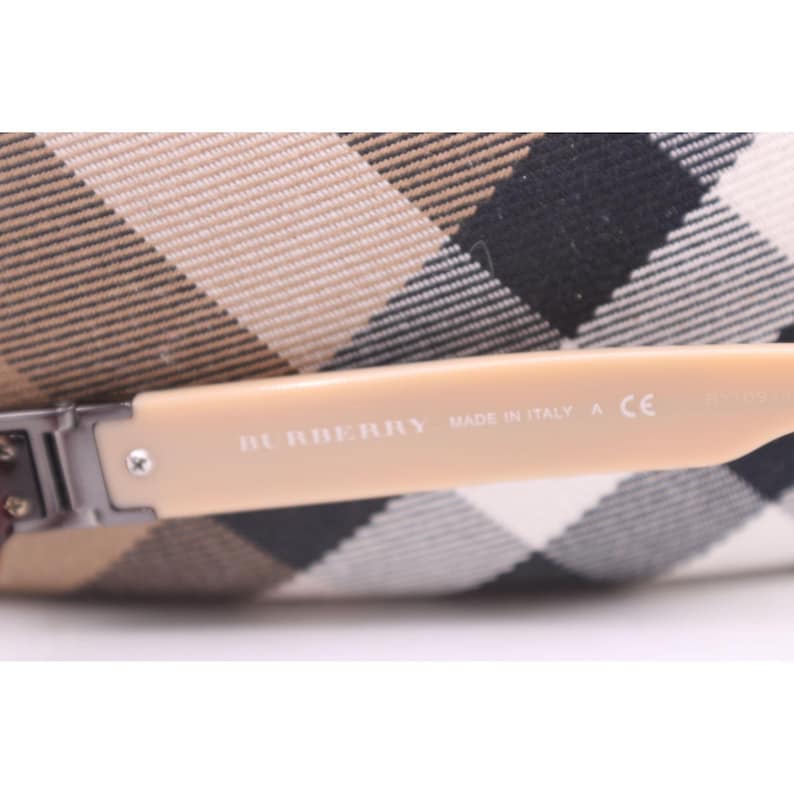 May include: Close-up of a pair of sunglasses with a light beige arm. The text "BURBERRY MADE IN ITALY A CE" is printed on the arm. The background features a beige and black plaid pattern.
