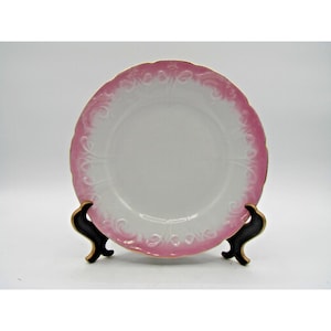 May include: A white porcelain plate with a pink floral border and gold trim. The plate is displayed on a black stand against a white background. The plate has a scalloped edge and embossed floral details.
