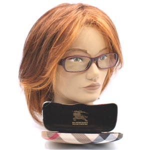 May include: A pair of rectangular, burgundy-framed eyeglasses on a mannequin head. The glasses are in front of an open case with a plaid pattern and the Burberry logo. The mannequin has auburn hair.