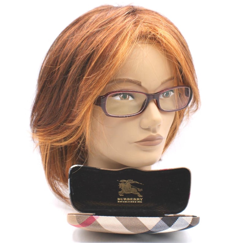 May include: A pair of rectangular, burgundy-framed eyeglasses on a mannequin head. The glasses are in front of an open case with a plaid pattern and the Burberry logo. The mannequin has auburn hair.