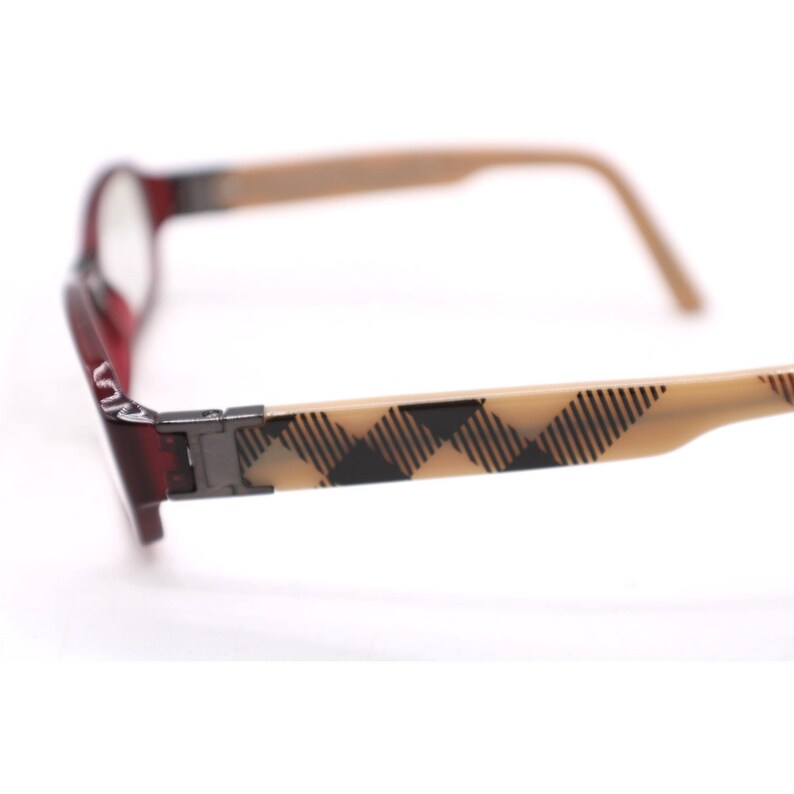 May include: A pair of eyeglasses with a red frame and tan arms. The arms feature a black and tan plaid pattern. The lenses are clear, and the glasses are set against a white background.