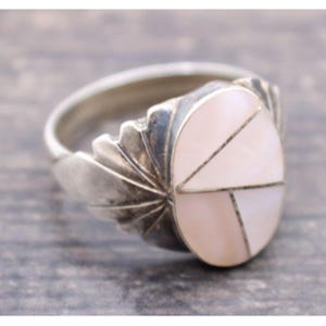 May include: A silver ring featuring an oval-shaped inlay of iridescent white shell divided into three geometric segments. The metal band displays a fan-like, pleated design extending from the sides of the central oval setting.