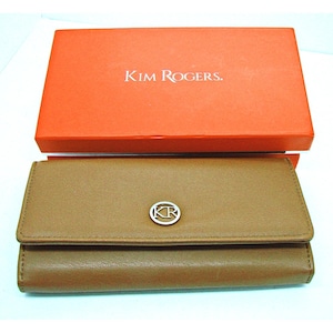 May include: A tan rectangular wallet with a fold-over flap and a silver circular logo featuring the letters KR. The wallet is positioned in front of an orange rectangular box with the white text KIM ROGERS. printed on the top surface.
