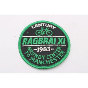 May include: A circular embroidered patch with a green border and black background. It features a green bicycle icon, the text CENTURY, RAGBRAI XI, 1983, and GRUNDY CENTER TO MANCHESTER in white and green embroidery thread.