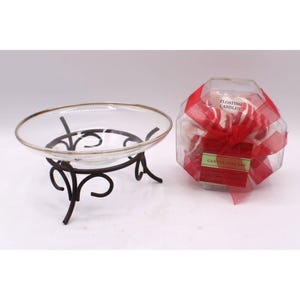 May include: A clear glass bowl with a metallic rim resting on a dark brown metal stand with scrollwork legs, positioned next to a hexagonal clear plastic box containing floating candles, wrapped in a red ribbon with a gift tag labeled Candle Gift Set.