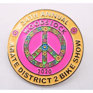 May include: A round, colorful pin with the text "34th Annual Woodstock Bike Show 2020." The pin features a peace sign in the center, surrounded by flowers and a yellow border. The text "ABATE DISTRICT 2 BIKE SHOW" is also visible.