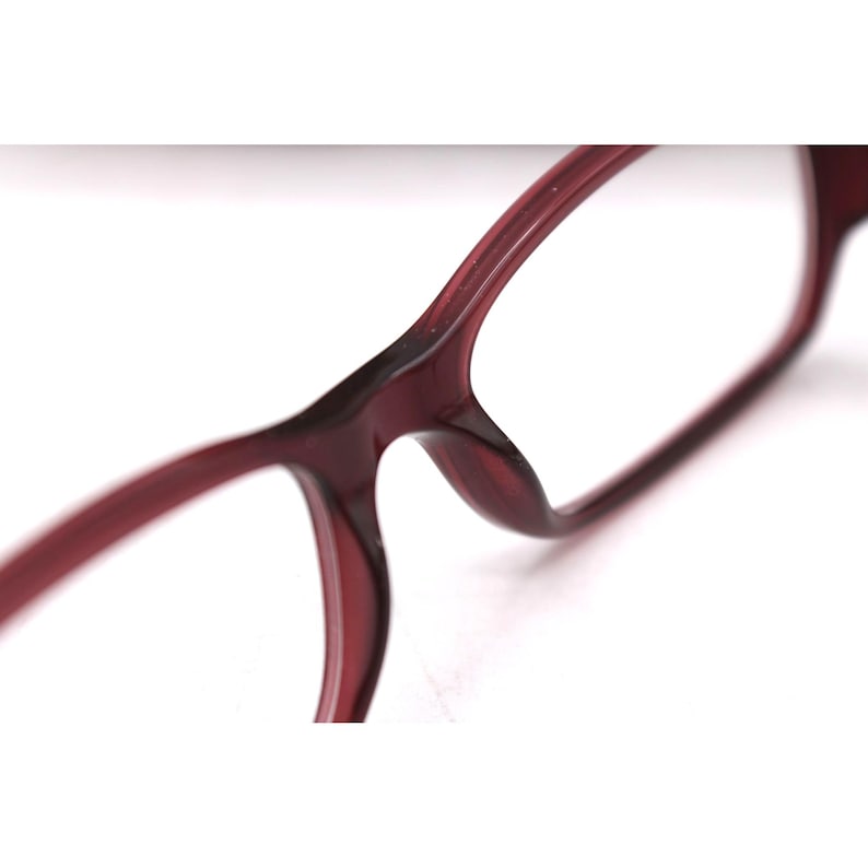 May include: Close-up of burgundy-colored eyeglasses. The frames have a rounded rectangular shape and are made of a smooth, glossy material. The design features a bridge and temples, with a focus on the sleek, modern aesthetic.