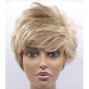 May include: A short, layered blonde wig with a slightly tousled style. The wig is displayed on a mannequin head with realistic features, including blue-gray eyes and full lips. The hair color is a light, warm blonde.