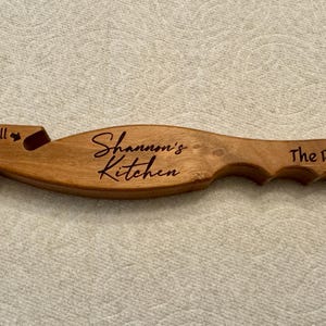Peut inclure: Un ustensile de cuisine en bois de couleur marron clair, de forme incurvée. L'outil a une extrémité fourchue avec les instructions "push" et "pull". Le texte "Shannon's Kitchen" et "The Rack Hooker" est gravé.