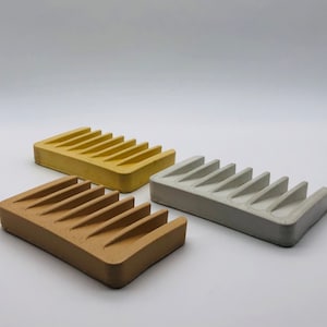 May include: Three soap dishes made of concrete in different colours: yellow, brown, and grey. Each dish has a rectangular shape with seven ridges.