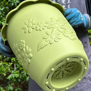 May include: A light green plastic flower pot with embossed floral designs. The pot has a scalloped rim and drainage holes at the bottom. The pot is held by a person wearing blue gloves.