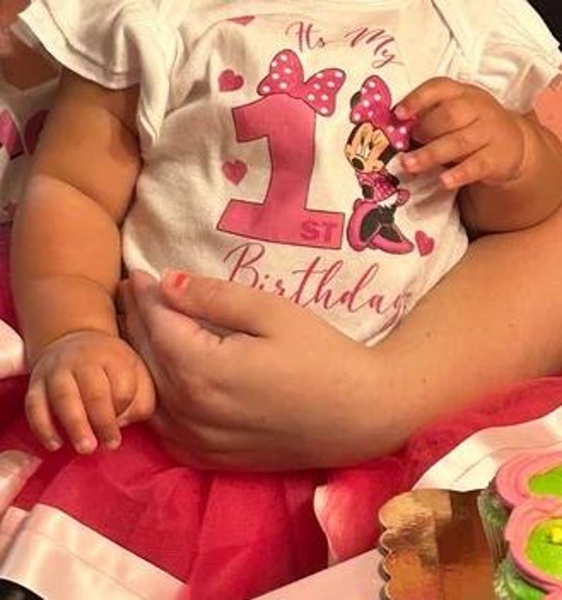 Op de afbeelding: Een babymeisje draagt een wit rompertje met een roze strik en de tekst "It's My 1st Birthday" en een Minnie Mouse-afbeelding. De baby draagt een roze tutu en heeft roze nagellak.