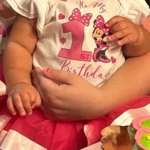 May include: A baby girl wearing a white onesie with a pink bow and the text "It's My 1st Birthday" and a Minnie Mouse graphic. The baby is wearing a pink tutu and has pink nail polish.