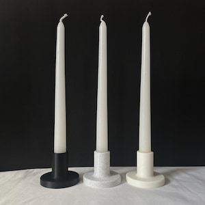 May include: Three white taper candles in different coloured candle holders. The candle holders are black, white and grey.