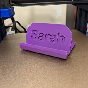 May include: A purple 3D printed phone stand with the name "Sarah" printed on the front.