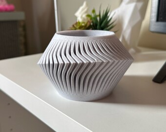 May include: A light grey, textured vase with a unique, geometric design. The vase features a series of vertical, wave-like cuts that create a textured surface. The vase is on a white surface.