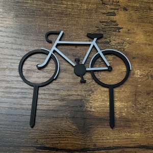 May include: Black metal bicycle silhouette cake topper with two black sticks. The bicycle is silver and has a black seat.