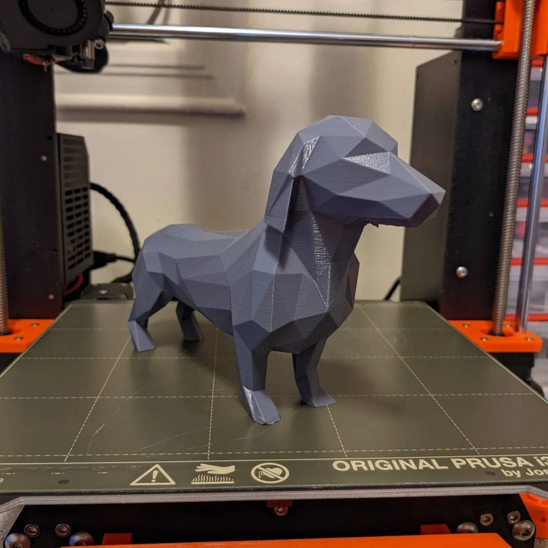 Puede incluir: Un modelo de perro salchicha impreso en 3D de color gris. El modelo est&aacute; de pie sobre una superficie texturizada negra y gris. El modelo est&aacute; hecho de pl&aacute;stico y tiene un estilo de bajo pol&iacute;gono.