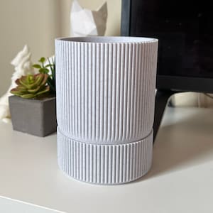 Könnte beinhalten: Ein weißer Keramik-Pflanztopf mit geripptem Design. Der Topf hat eine strukturierte Oberfläche und einen leicht erhöhten Boden.