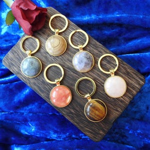 May include: Six circular keychains with gold-toned metal rings and settings, each featuring a different polished gemstone or cabochon. The stones include variations of tan, gray, purple, coral, tiger's eye, and pale pink, arranged on a dark wood board.