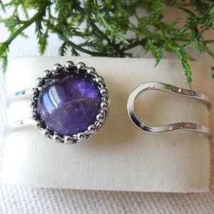 May include: A silver-toned cuff bracelet featuring a round, polished purple amethyst cabochon set in a beaded metal bezel. The bracelet has an open design with a smooth, curved band that tapers toward the ends, showcasing a metallic finish.
