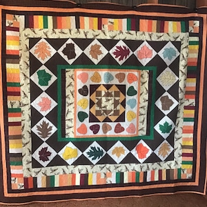 May include: A brown, green, and orange quilt with a pattern of leaves and hearts. The quilt is made with a variety of fabrics and has a quilted design.