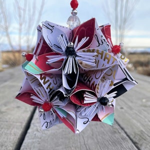 May include: A red, white, and black paper ornament with a white string hanging from it. The ornament is made of folded paper and has a starburst shape. It is decorated with black and white paper flowers and red beads.