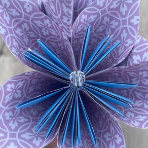 May include: A close-up of a handmade paper flower. The flower has purple petals with a white floral pattern and blue accents. A clear gem is in the center. The flower is a decorative craft item.