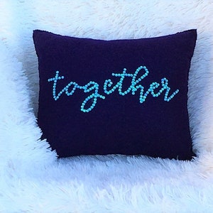 May include: A square, dark purple pillow with the word "together" embroidered in cursive using small, light blue beads. The pillow is resting on a fluffy, white surface.