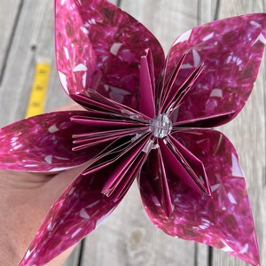 May include: A handmade paper flower with a vibrant magenta and white pattern. The flower has six large petals and a central cluster of smaller, folded petals. A clear, faceted bead is at the center. The flower is held against a wooden background.
