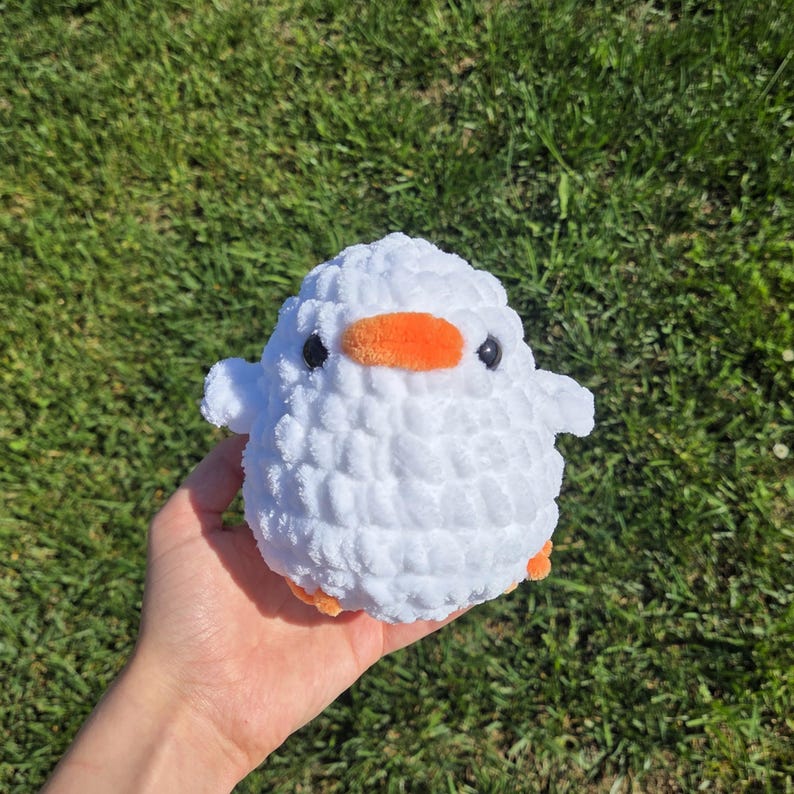 May include: A handmade white crochet duck with an orange beak and feet, and black eyes. The plush toy is round and fluffy, with small wings on either side. The duck is held in a hand against a background of green grass.
