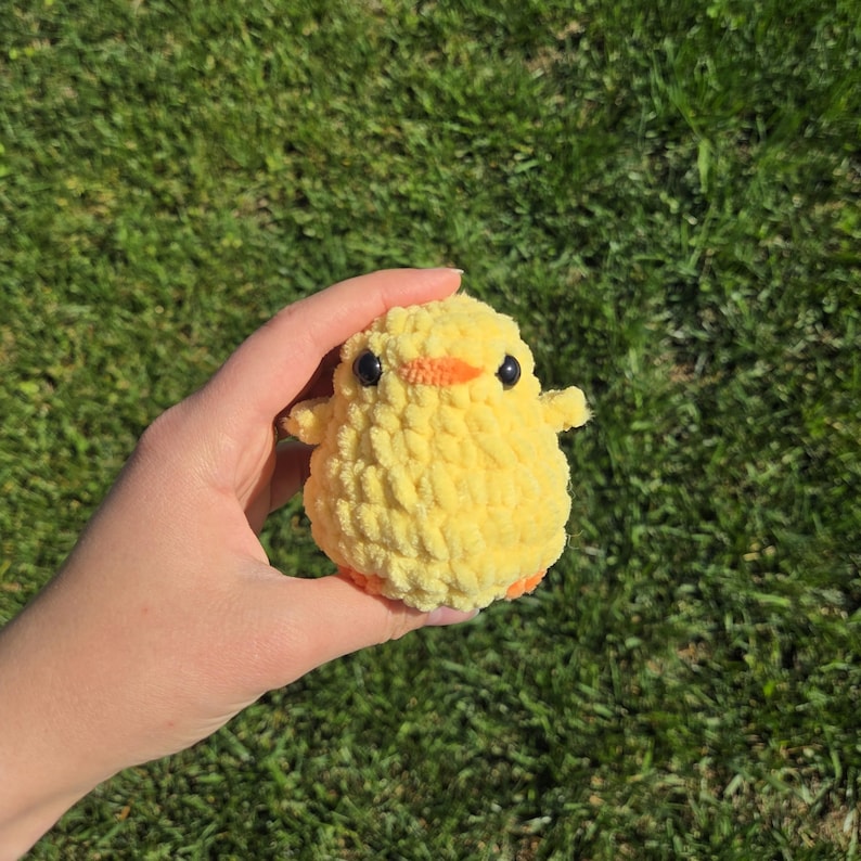 May include: A small, yellow crocheted chick with black eyes and an orange beak and feet. The chick has a fluffy texture and small wings. The chick is held in a hand against a background of green grass.