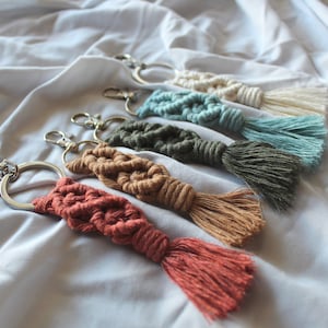 May include: A collection of macrame keychains in various colors, including coral, tan, olive green, teal, and cream. Each keychain features a woven knot design and a matching tassel, attached to a silver-tone metal ring and clasp.