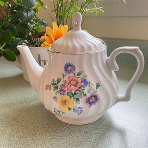 May include: White teapot with a floral design featuring pink, yellow, purple, and blue flowers. The teapot has a gold trim around the lid and spout.