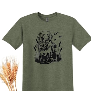 May include: Heather Military Green t-shirt featuring a black graphic of a dog holding a duck in its mouth, with cattails and flying birds. The shirt has short sleeves and a crew neck. The text "Heather Military Green" is at the bottom right.