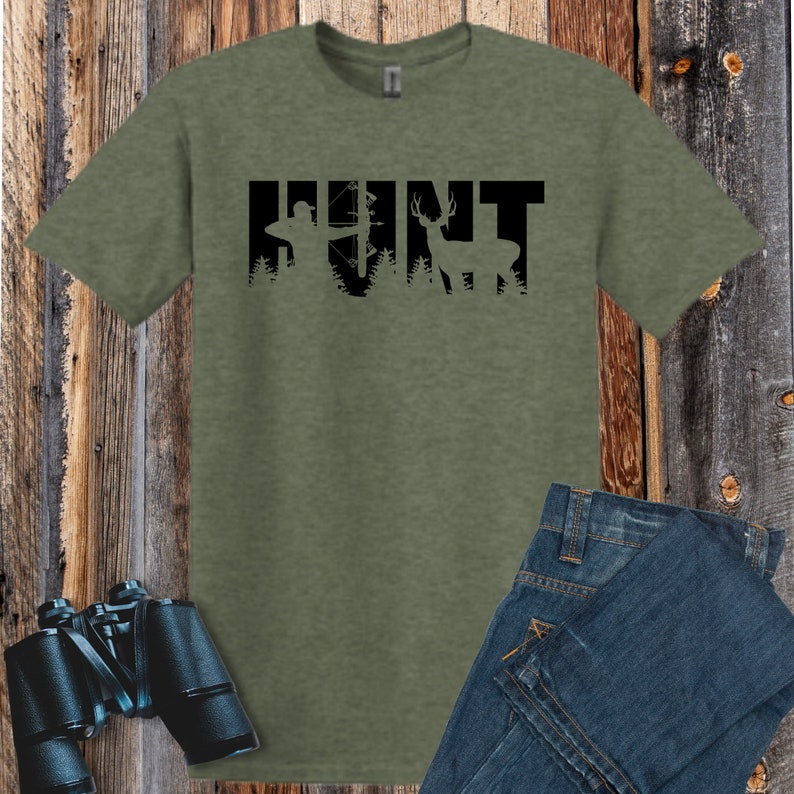Puede incluir: Una camiseta verde con la palabra "HUNT" en letras negras. Las letras est&aacute;n formadas por siluetas de un cazador con arco y flecha y un ciervo en el bosque.