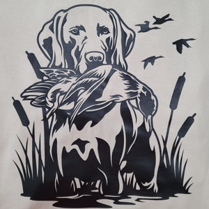 May include: A black and white graphic of a Labrador Retriever holding a duck in its mouth. The dog is surrounded by cattails and flying ducks, set against a light beige background. The design evokes a hunting or outdoor theme.