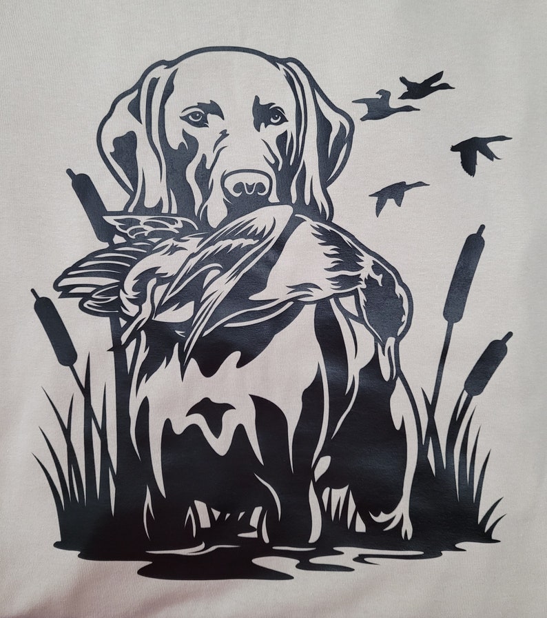 May include: A black and white graphic of a Labrador Retriever holding a duck in its mouth. The dog is surrounded by cattails and flying ducks, set against a light beige background. The design evokes a hunting or outdoor theme.