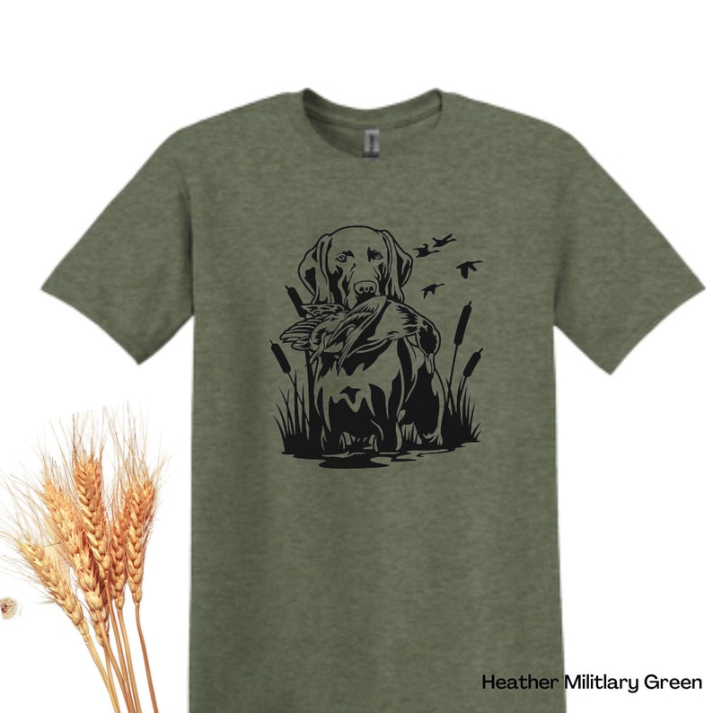May include: Heather Military Green t-shirt featuring a black graphic of a dog holding a duck in its mouth, with cattails and flying birds. The shirt has short sleeves and a crew neck. The text "Heather Military Green" is at the bottom right.