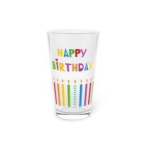 May include: A clear glass with the words "HAPPY BIRTHDAY" in colorful letters. The glass is decorated with a row of colorful candles along the bottom. A small red heart is between the words "HAPPY" and "BIRTHDAY".