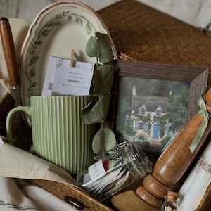 May include: A curated gift basket with a green mug, a framed watercolor painting of a house, and a small jar of tea bags. Includes a decorative plate, a wooden rolling pin, and a small bottle of matches. The basket is lined with a white cloth.