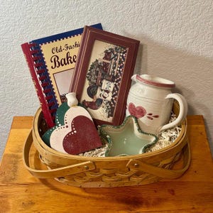 May include: A decorative basket filled with various kitchen-themed items. Includes a cookbook, framed picture, heart-shaped trivets, a star-shaped dish, and a ceramic pitcher. The cookbook reads "Old-Fashi Bake".