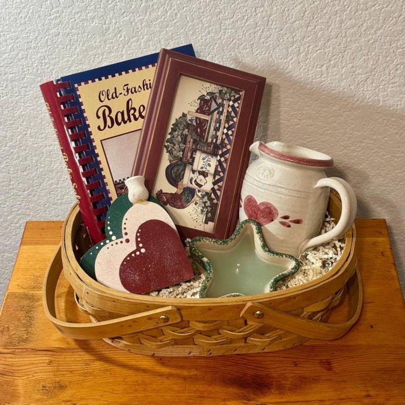 May include: A decorative basket filled with various kitchen-themed items. Includes a cookbook, framed picture, heart-shaped trivets, a star-shaped dish, and a ceramic pitcher. The cookbook reads "Old-Fashi Bake".