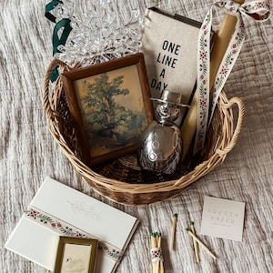 May include: A woven basket filled with decorative items. Includes a framed landscape painting, a journal with "ONE LINE A DAY" text, a snowman-shaped perfume bottle, a candle with a floral ribbon, a snowflake ornament, and stationery with a small framed picture.